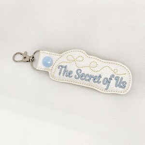 May include: White leather keychain with a blue button and the text "The Secret of Us" embroidered in blue thread.