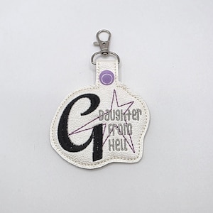 May include: A white, textured, irregular-shaped keychain featuring a large black embroidered letter G, a purple star outline, and the embroidered text Daughter from Hell. It includes a silver metal lobster clasp and a small purple snap button.