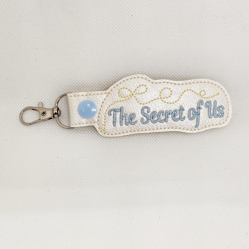 May include: White leather keychain with a blue button and the text "The Secret of Us" embroidered in blue thread.