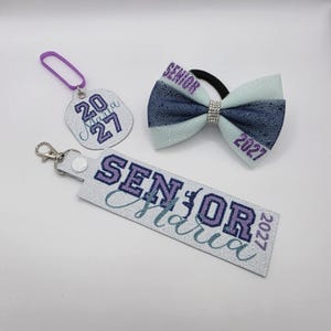 May include: A silver glitter keyring with the text SENIOR Maria 2027, a round silver glitter keyring with the text 20 27 and Maria, and a hair bow with a dark blue and light blue fabric, a rhinestone centre, and the text SENIOR 2027.