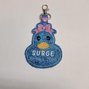Pu&ograve; includere: Un portachiavi a forma di anatra blu glitterata con un fiocco rosa, il testo "SURGE Sienna 2025" e un ciondolo a forma di cuore rosa.