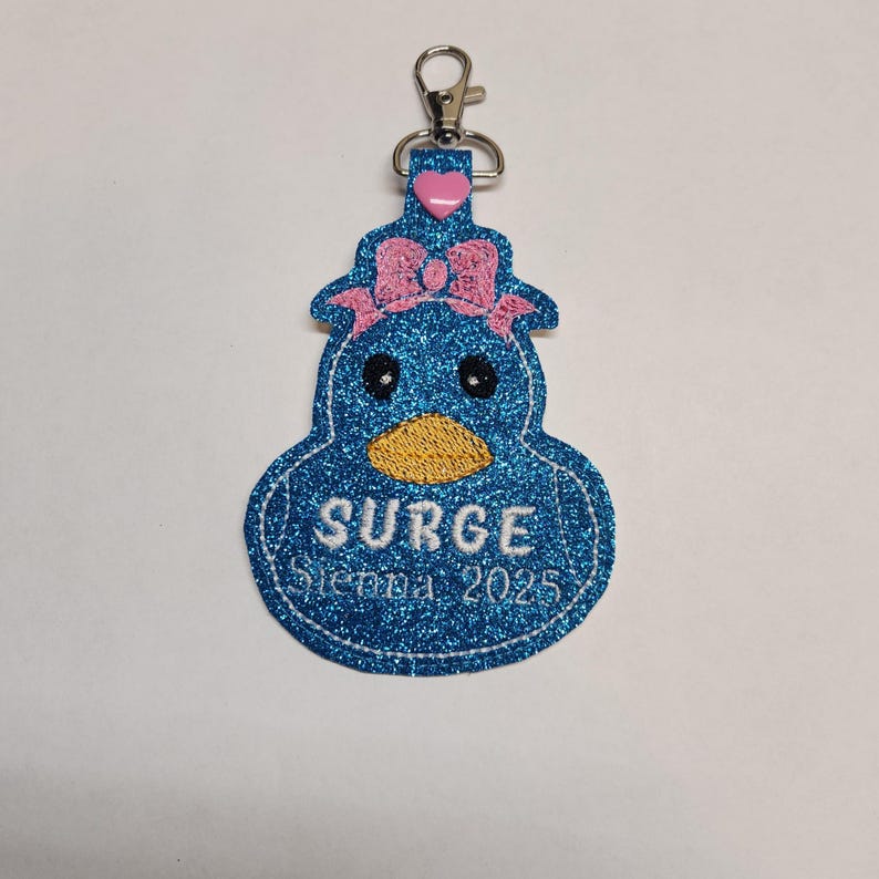 Pu&ograve; includere: Un portachiavi a forma di anatra blu glitterata con un fiocco rosa, il testo "SURGE Sienna 2025" e un ciondolo a forma di cuore rosa.