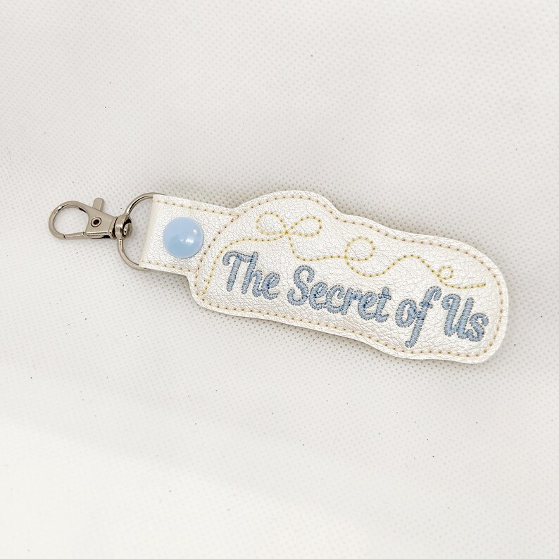 May include: White leather keychain with a blue button and the text "The Secret of Us" embroidered in blue thread.