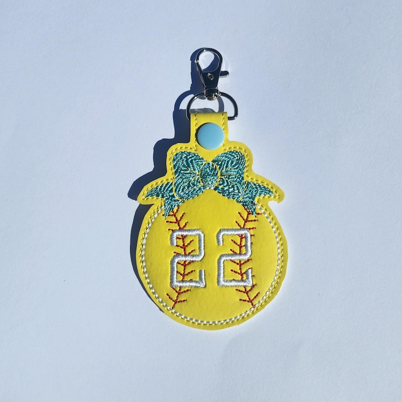 Puede incluir: Llavero de softball amarillo con un lazo azul y el n&uacute;mero 22 bordado.