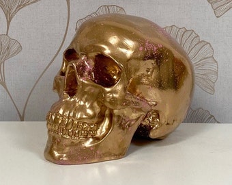 Pode incluir: Uma caveira dourada com detalhes em rosa. A caveira é feita de um material liso e brilhante e tem um design detalhado.