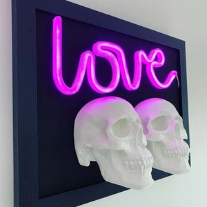 May include: A framed artwork featuring a neon pink "love" sign and two white skull sculptures. The artwork has a black background and a dark blue frame.