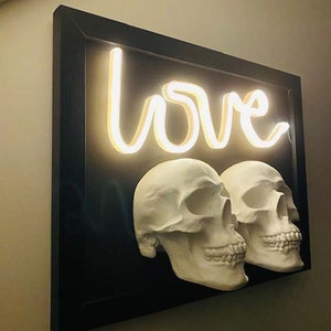 May include: A black framed artwork featuring a neon sign that reads "love" and two white plaster skulls.