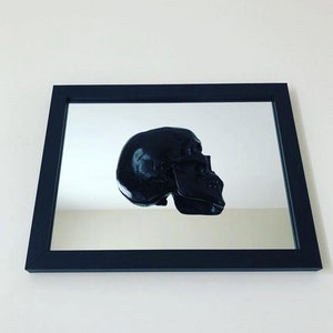 May include: A black skull sculpture reflected in a black framed mirror.