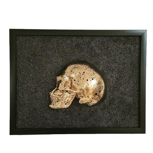 May include: A gold-coloured skull sculpture is framed against a black, sparkly background. The skull is positioned in profile, with the left side of the skull facing the viewer.