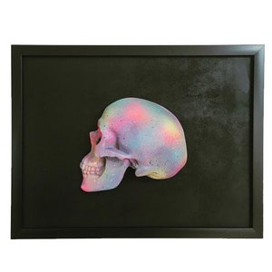 May include: A colourful, pink, blue and purple skull sculpture in a black frame. The skull is painted with a galaxy-like design.