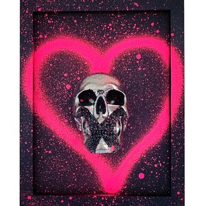 May include: A silver skull with a black X over its mouth is framed in a black frame with a pink heart spray painted on a black background.