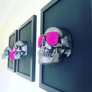 May include: Three framed silver skull sculptures with pink rhinestone sunglasses. The skulls are mounted on black backgrounds and framed in black.