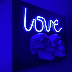 May include: A black framed wall art piece with a blue neon sign that says "love" and two white skulls below the sign.