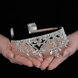 K&ouml;nnte beinhalten: Eine silberne Tiara mit einem zarten, filigranen Design, das mit klaren Strasssteinen besetzt ist. Die Tiara hat einen Kamm auf der R&uuml;ckseite, um sie leicht im Haar zu befestigen.