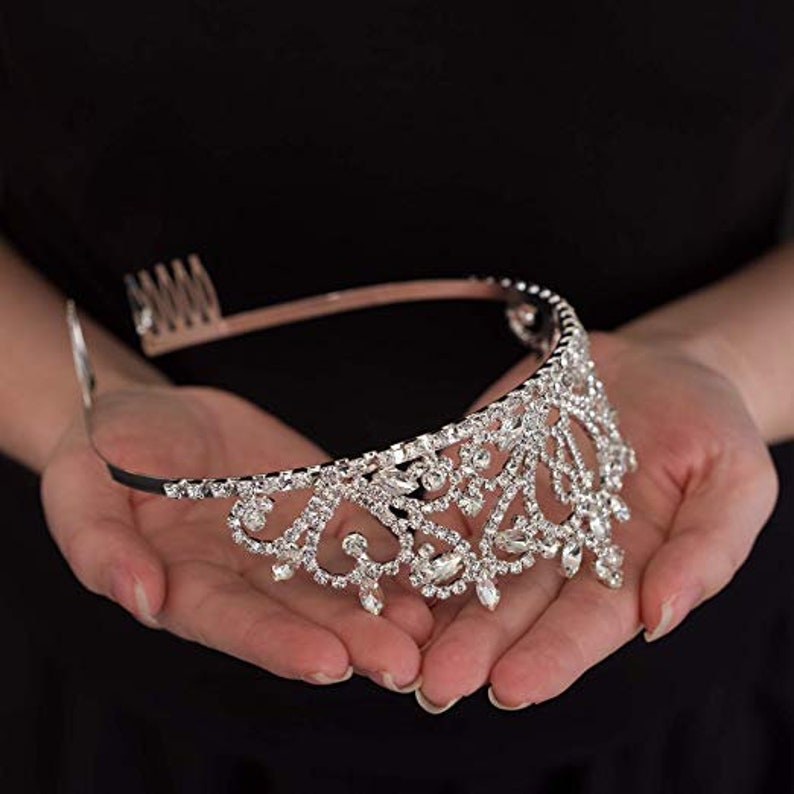 May include: A silver tiara with a delicate, intricate design featuring clear rhinestones. The tiara has a flexible band and a comb for secure placement.