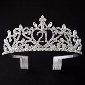 May include: Silver tiara with rhinestones and a heart-shaped design featuring the number 21. The tiara has two combs on the back for securing it in hair.