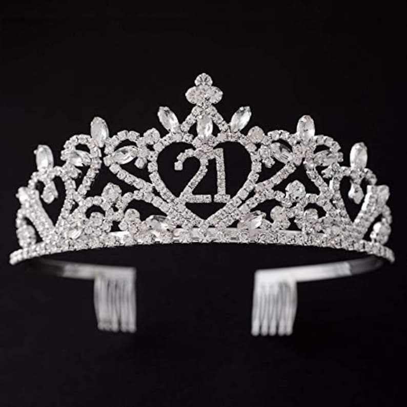 May include: Silver tiara with rhinestones and a heart-shaped design featuring the number 21. The tiara has two combs on the back for securing it in hair.