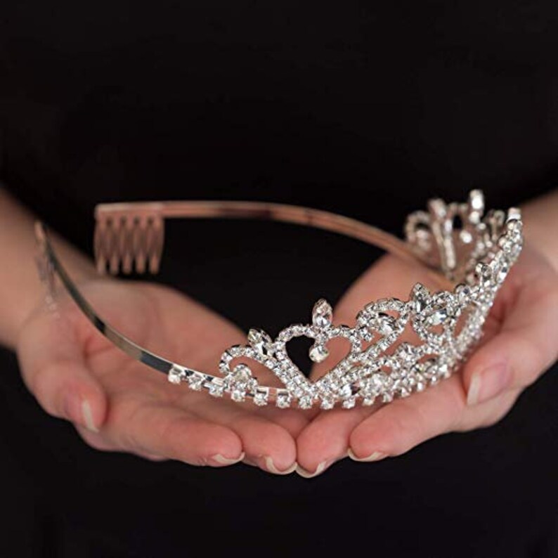 May include: A silver tiara with a heart-shaped design and clear rhinestones. The tiara is held in the hands of a person.