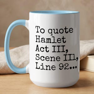 Op de afbeelding: Witte keramische mok met een lichtblauw handvat en interieur. De mok bevat de quote "To quote Hamlet Act III, Scene III, Line 92..." in zwarte tekst. De mok staat op een houten oppervlak.