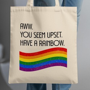 Op de afbeelding: Een crèmekleurige draagtas met de tekst "Aww. You seem upset. Have a rainbow." en een regenboogafbeelding. De tas heeft twee handvatten. De tas wordt door een persoon vastgehouden.