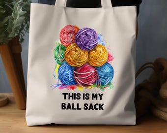 May include: A white tote bag featuring a watercolor illustration of colorful yarn balls in red, purple, green, yellow, and blue. The text "THIS IS MY BALL SACK" is printed in black below the yarn balls.