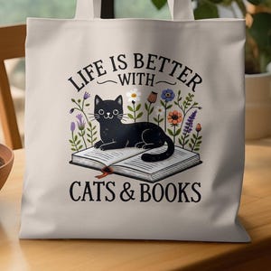 May include: Cream-colored tote bag featuring a black cat lying on an open book, surrounded by colorful flowers. The text "LIFE IS BETTER WITH CATS & BOOKS" is printed above and below the cat illustration.