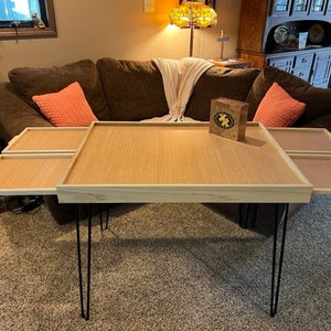 May include: A wooden coffee table with four removable trays. The table has a light wood finish and black hairpin legs. The trays are made of a darker wood and have a smooth surface.
