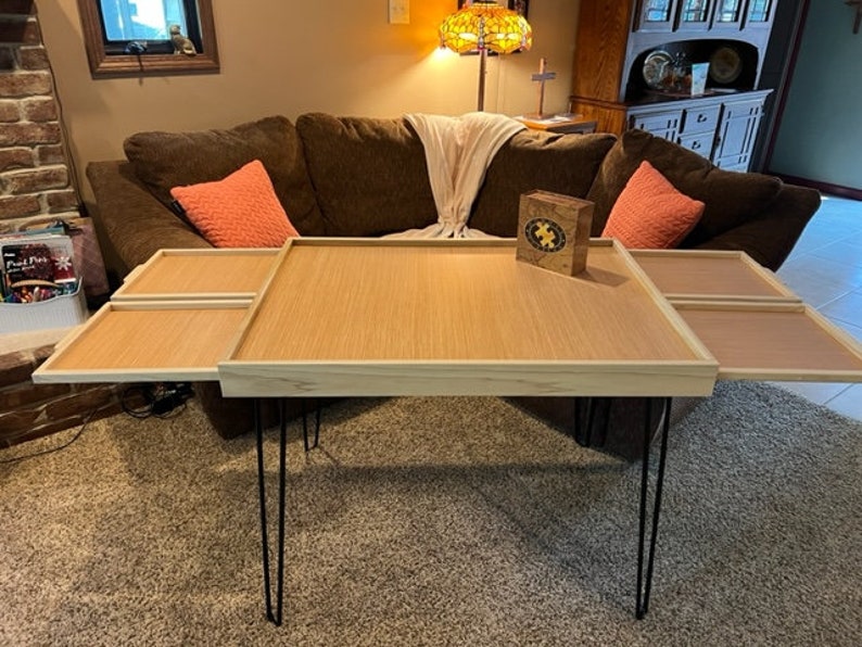 May include: A wooden coffee table with four removable trays. The table has a light wood finish and black hairpin legs. The trays are made of a darker wood and have a smooth surface.