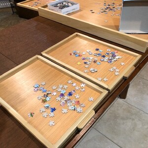 May include: Two wooden puzzle trays with a light brown finish, each filled with colourful puzzle pieces. The trays are on a dark brown wooden table.