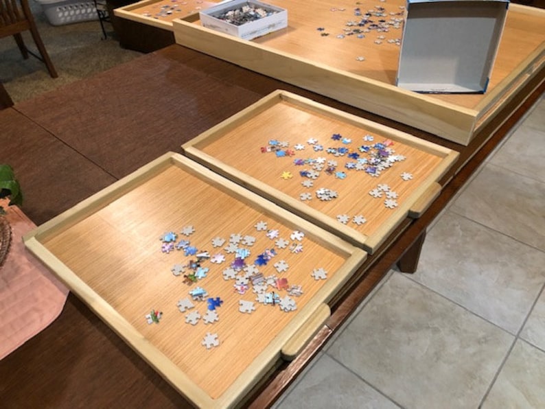 May include: Two wooden puzzle trays with a light brown finish, each filled with colourful puzzle pieces. The trays are on a dark brown wooden table.