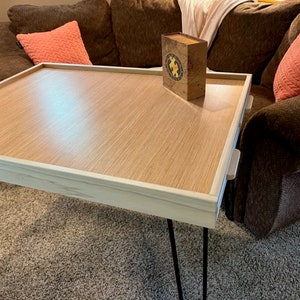 May include: A light wood tray table with a white border and black hairpin legs. The table is sitting on a brown carpet and has a small box on top of it.