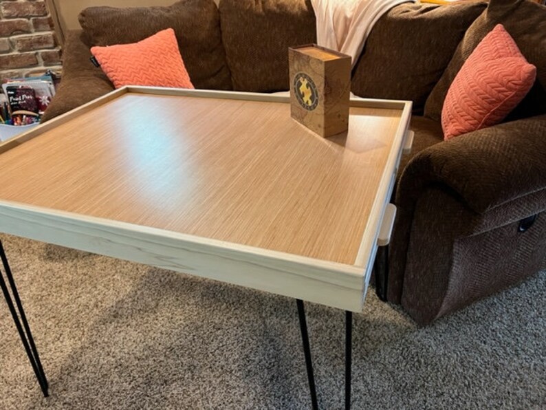 May include: A light wood tray table with a white border and black hairpin legs. The table is sitting on a brown carpet and has a small box on top of it.