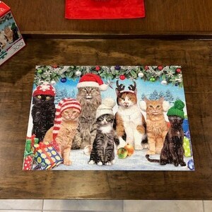 May include: A completed 300-piece jigsaw puzzle depicting a group of cats wearing festive holiday hats and jumpers. The cats are surrounded by Christmas decorations, including baubles, garlands, and presents.