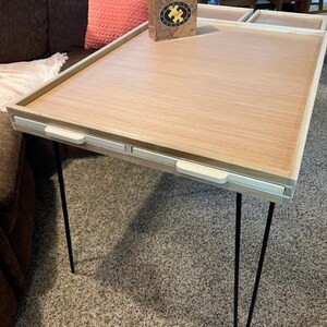 May include: A wooden table with a light wood top and white trim. The table has three black hairpin legs and three white drawers with white handles. The table is sitting on a brown carpet.