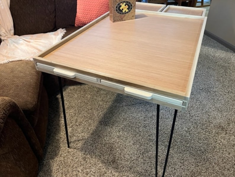 May include: A wooden table with a light wood top and white trim. The table has three black hairpin legs and three white drawers with white handles. The table is sitting on a brown carpet.
