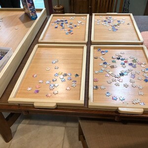 May include: Five wooden puzzle trays with a light wood finish and a raised edge. Each tray has a different number of puzzle pieces scattered on the surface. The pieces are colourful and have a variety of shapes.