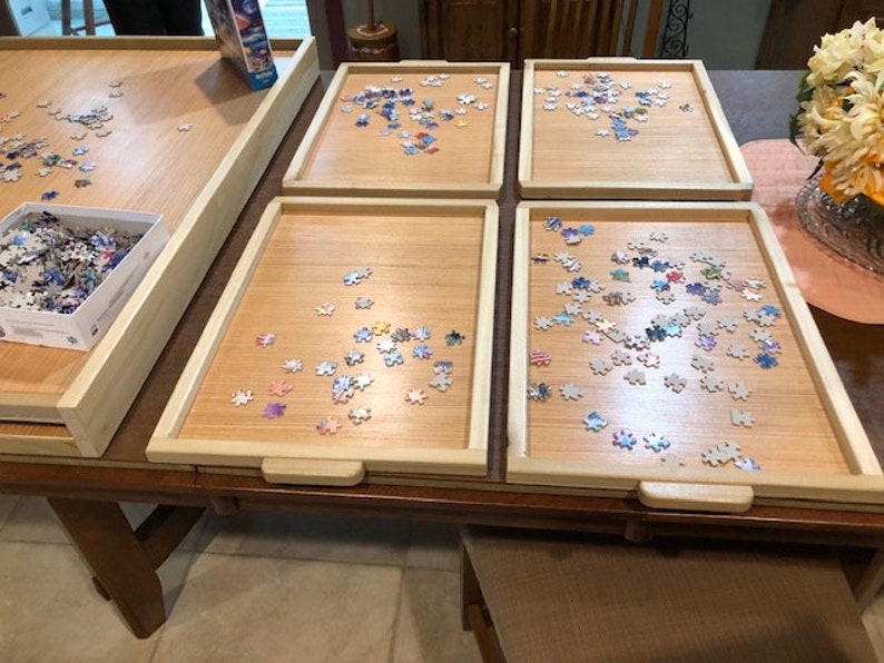 May include: Five wooden puzzle trays with a light wood finish and a raised edge. Each tray has a different number of puzzle pieces scattered on the surface. The pieces are colourful and have a variety of shapes.