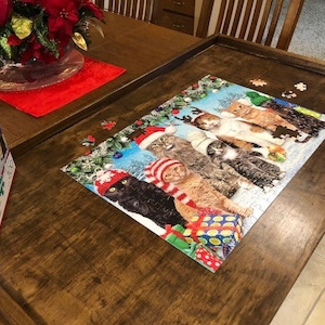May include: A completed jigsaw puzzle featuring a group of cats wearing festive winter hats and holding gifts. The puzzle is set on a wooden table with a red tablecloth and a vase of red poinsettias.