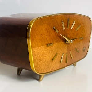 May include: A vintage wooden clock with a curved, dark brown top and a light brown face. The clock face has gold-colored hands and markers. The clock is rectangular with gold trim and sits on three gold-colored legs.