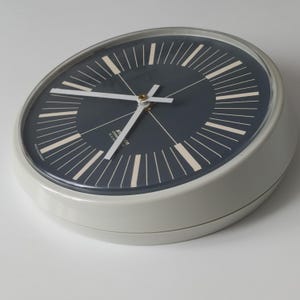May include: A round, gray wall clock with a dark blue face and white hands. The clock features a minimalist design with white markers and a gold-colored center. The clock's face has the text "STR" and "DDR" printed on it.