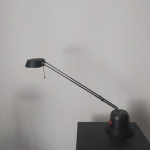Può includere: Una lampada da scrivania nera con testa e base circolari, collegate da due bracci dritti e argentati. La lampada ha un pulsante rosso sulla base e un cordino rosso. La lampada è su una superficie nera.