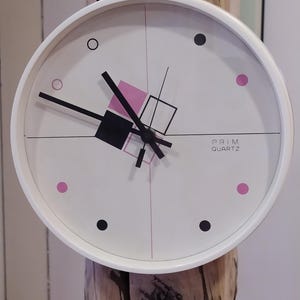 May include: White analog clock with black hands and pink and black geometric shapes on the face. The clock face also has black dots around the edge. The clock says "PRIM QUARTZ" at the bottom.