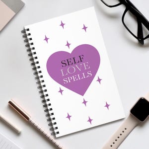 Puede incluir: Un cuaderno blanco con espiral con el texto "SELF LOVE SPELLS" dentro de un corazón morado. Estrellas moradas salpican el diseño. También se ven un bolígrafo, gafas y un reloj inteligente.
