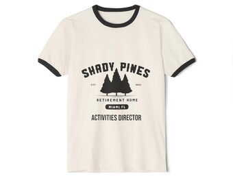 May include: Cream-coloured t-shirt with black trim around the collar and sleeves. The shirt features the text "SHADY PINES" above a graphic of three pine trees. Below the trees, the text reads "RETIREMENT HOME MIAMI FL ACTIVITIES DIRECTOR."