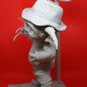 May include: A gray, unpainted figurine of a creature wearing a hat and holding a sign that says "SUR". The creature has large ears and claws. The figurine is standing on a gray base.