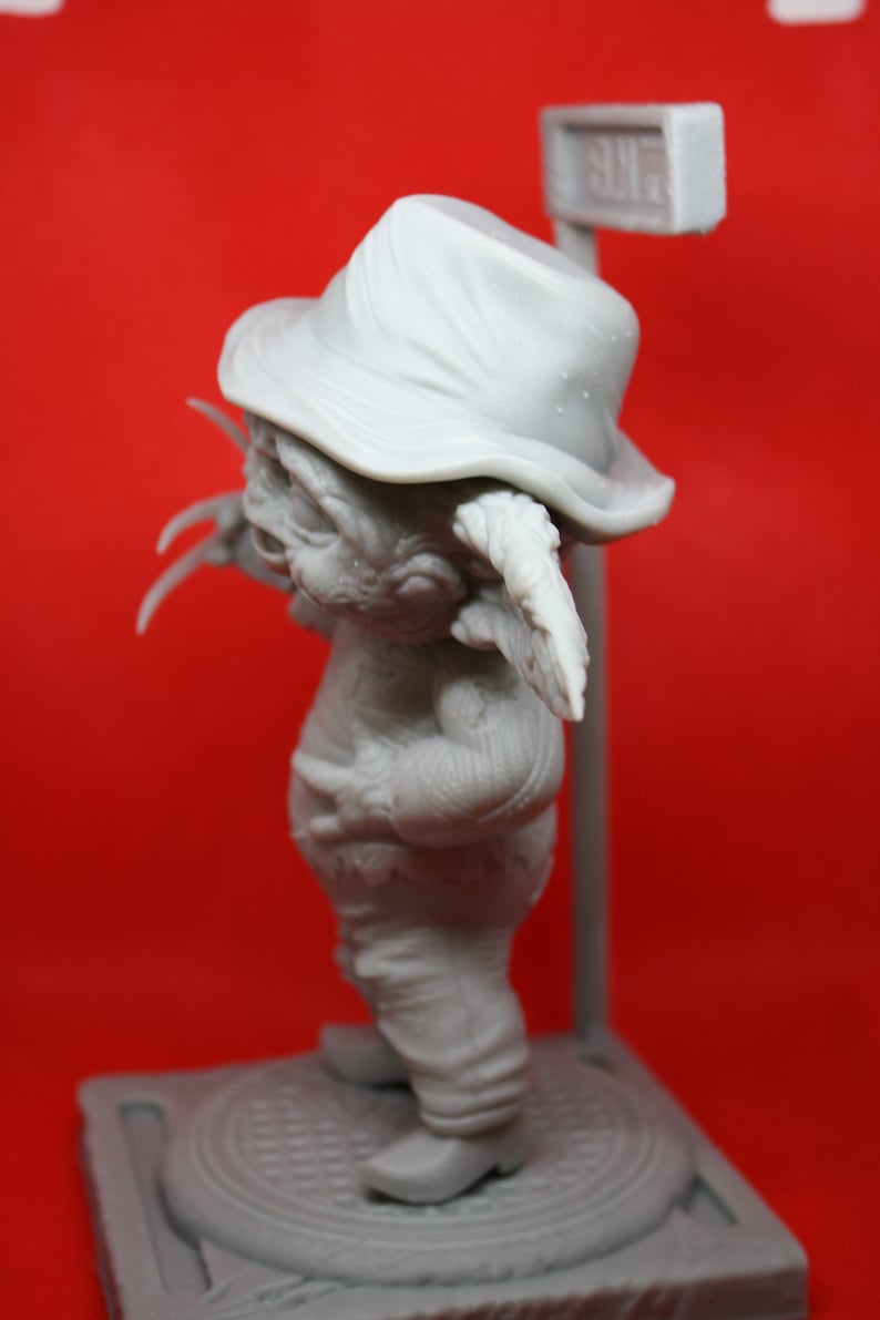 May include: A gray, unpainted figurine of a creature wearing a hat and holding a sign that says "SUR". The creature has large ears and claws. The figurine is standing on a gray base.