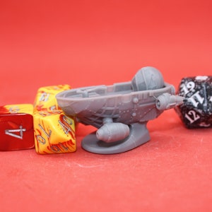 May include: A gray plastic miniature spaceship with a cockpit and a small engine on the bottom. The spaceship is surrounded by three multi-colored dice.