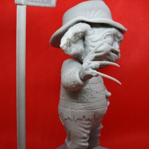 May include: A gray, 3D printed figurine of Freddy Krueger, the iconic horror character from the Nightmare on Elm Street franchise. The figurine is wearing a striped sweater and a hat, and is holding a glove with razor-sharp blades. The figurine is standing on a base with a sewer grate design and a street sign that reads "Elm St."
