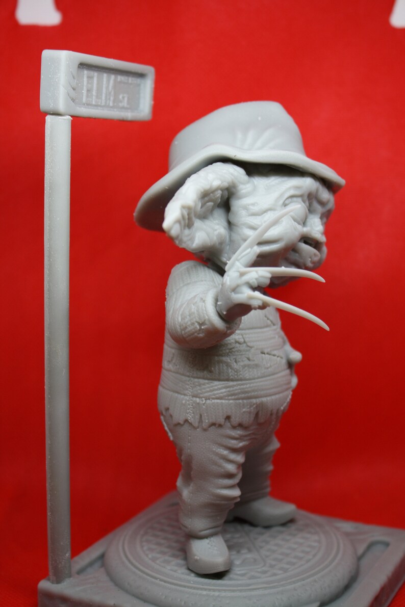 May include: A gray, 3D printed figurine of Freddy Krueger, the iconic horror character from the Nightmare on Elm Street franchise. The figurine is wearing a striped sweater and a hat, and is holding a glove with razor-sharp blades. The figurine is standing on a base with a sewer grate design and a street sign that reads "Elm St."