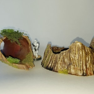 May include: A brown, hollowed-out tree trunk with green moss and a small figure standing in the opening. The tree trunk is split in half, with one half lying on its side and the other half standing upright. The tree trunk is made of a textured material that resembles wood.